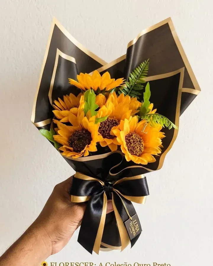 Girassóis artificiais com folhagens e embalagem preta com acabamento dourado.